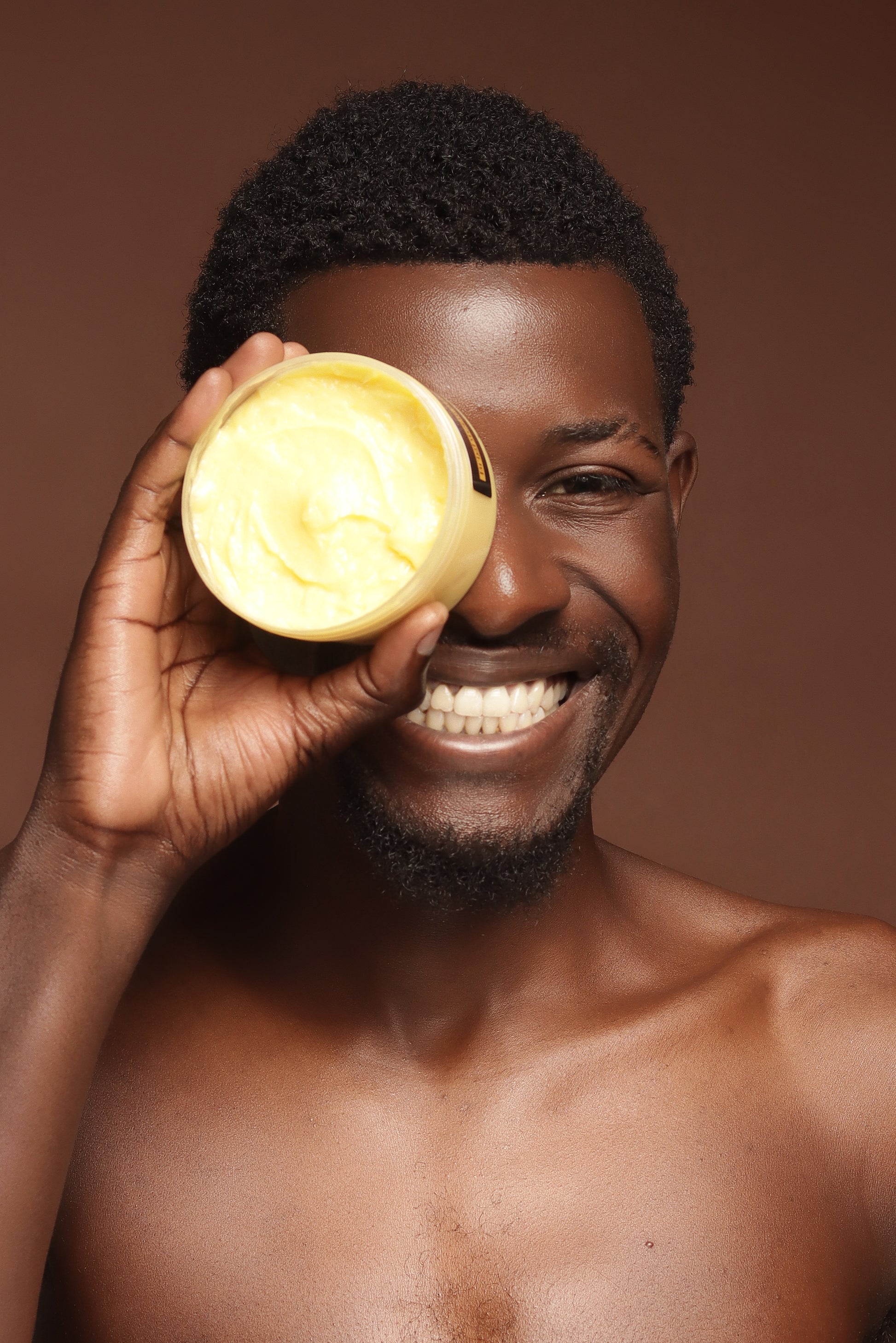 Man holding a Brightening Body Butter container against his eye on a brown background